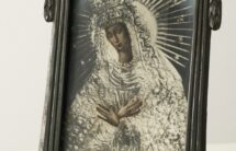Fotografia czarno-biała przedstawiającego Matkę Boską Ostrobramską. Postać kobiety widoczna od pasa w gorę, lekko zwrócona w lewą stronę. Srebrna szata, na głowie welon i dwie korony, jedna nad drugą. Wokół głowy nimb z długimi promieniami, pomiędzy nimi gwiazdki. Twarz łagodna, dłonie złożone na piersi. Twarz i dłonie lekko podbarwione na beżowo. Fotografia w metalowej stojącej ramce zdobionej ornamentami secesyjnymi.